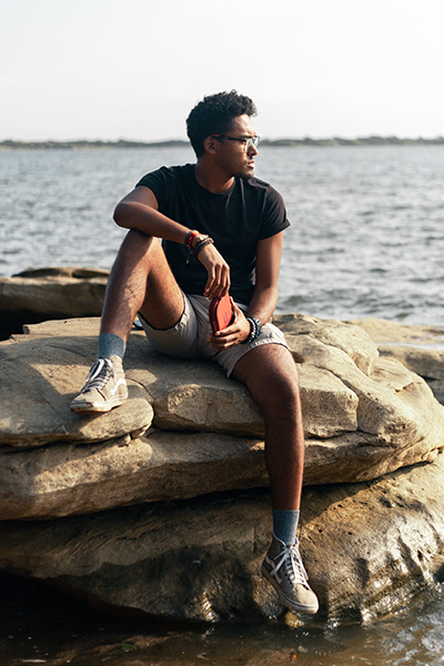 Man in a t-shirt and shorts sits on a rock formation near a beach during the sunny day