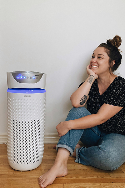 Smiling woman sits on wood floor next to a Homedics TotalClean® 5-in-1 UV-C Deluxe Large Room Air Purifier