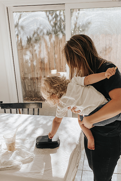 Woman in black jeans and t-shirt holds baby who reaches down to touch a toy on a table