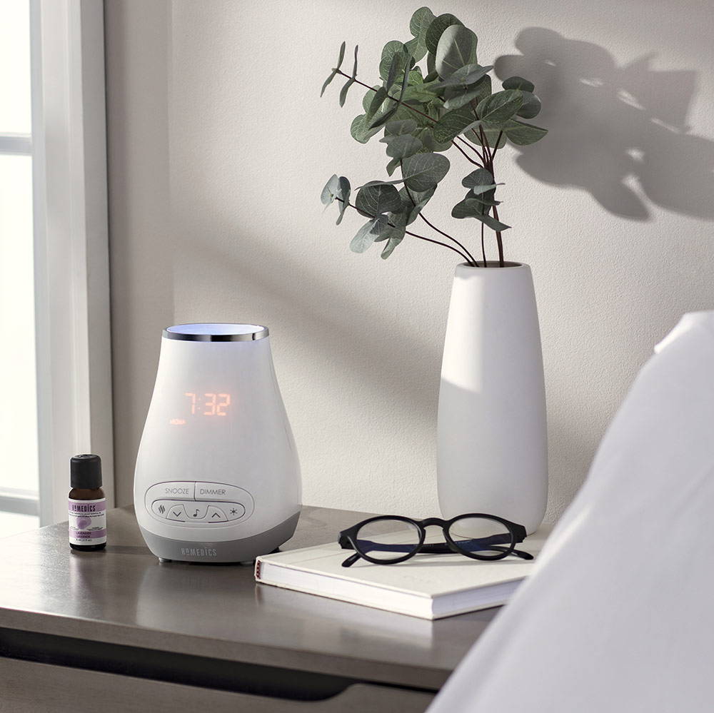 Homedics diffuser and essential oil bottle sit on a table next to a potted plant, eye glasses, and book.