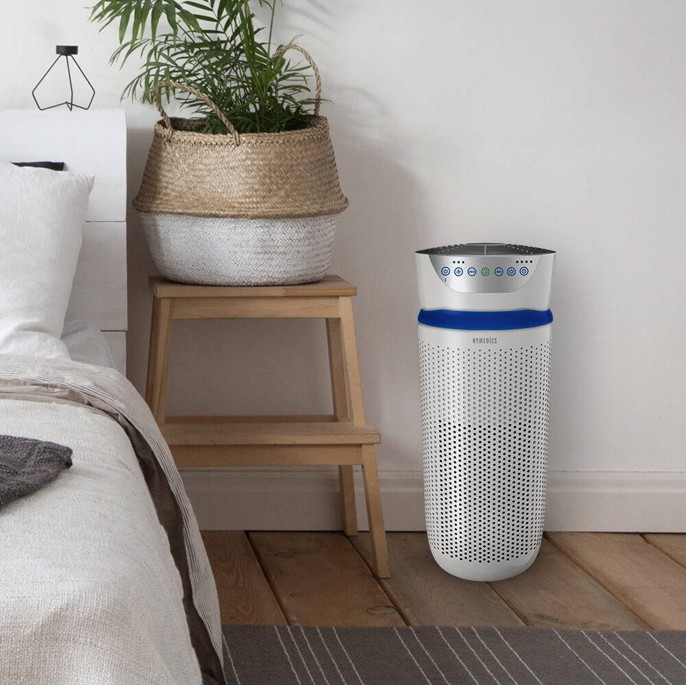 Homedics TotalClean air purifier in a brightly lit bedroom next to a bed.
