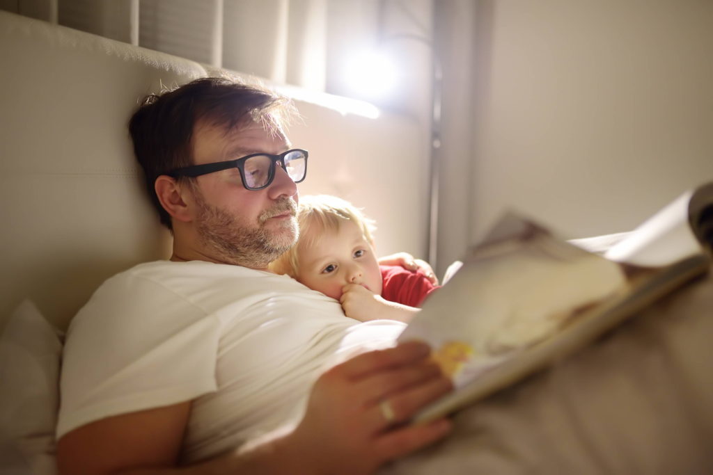 Man in white t-shirt lays in bed with small child and reads a bedtime story.