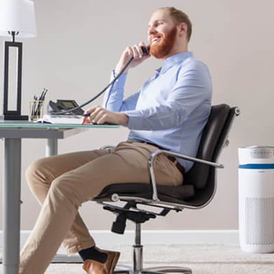 Man in business attire smiles while on the phone at office desk with Homedics air purifier in the background.