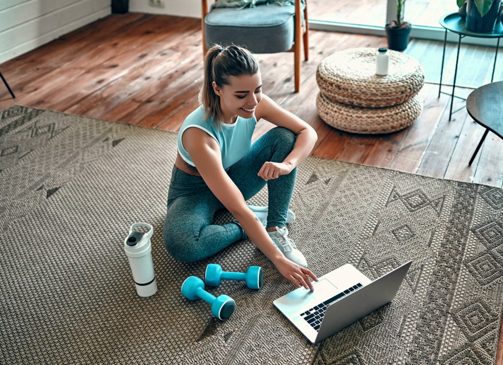 Woman in athletic clothing sits on the floor next to dumb bells while using laptop.