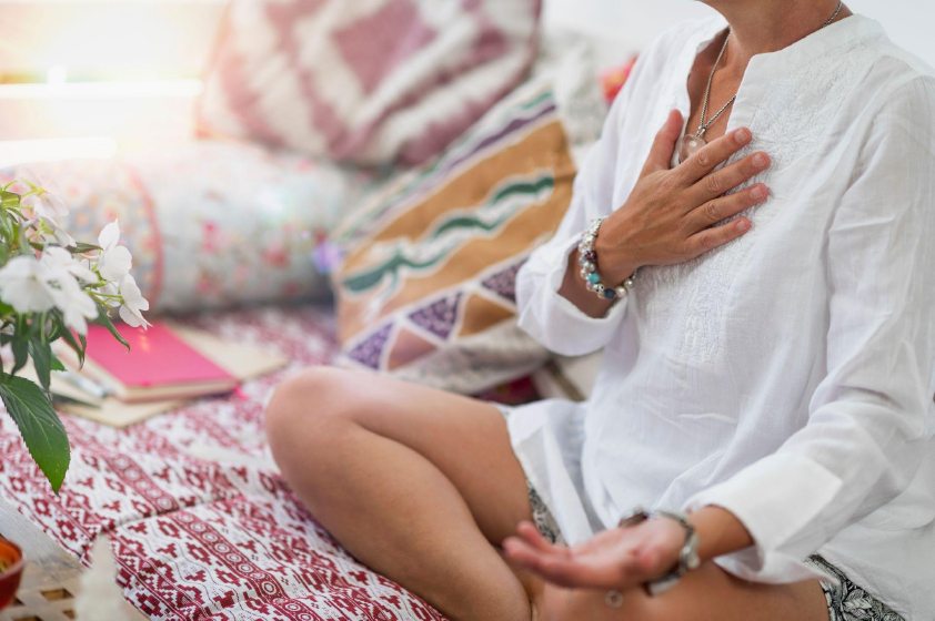 woman in white tunic sits on couch with legs folded and hand held to chest while meditating