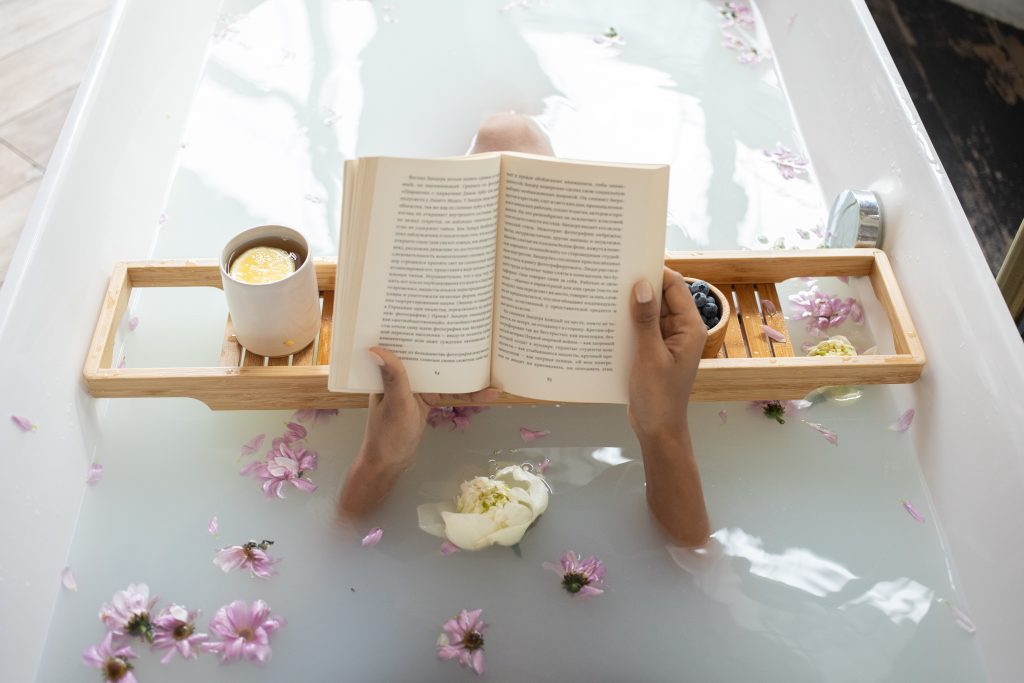 woman soaks in a hot bath with flowers while reading a book above tray with a cup of tea and blueberries