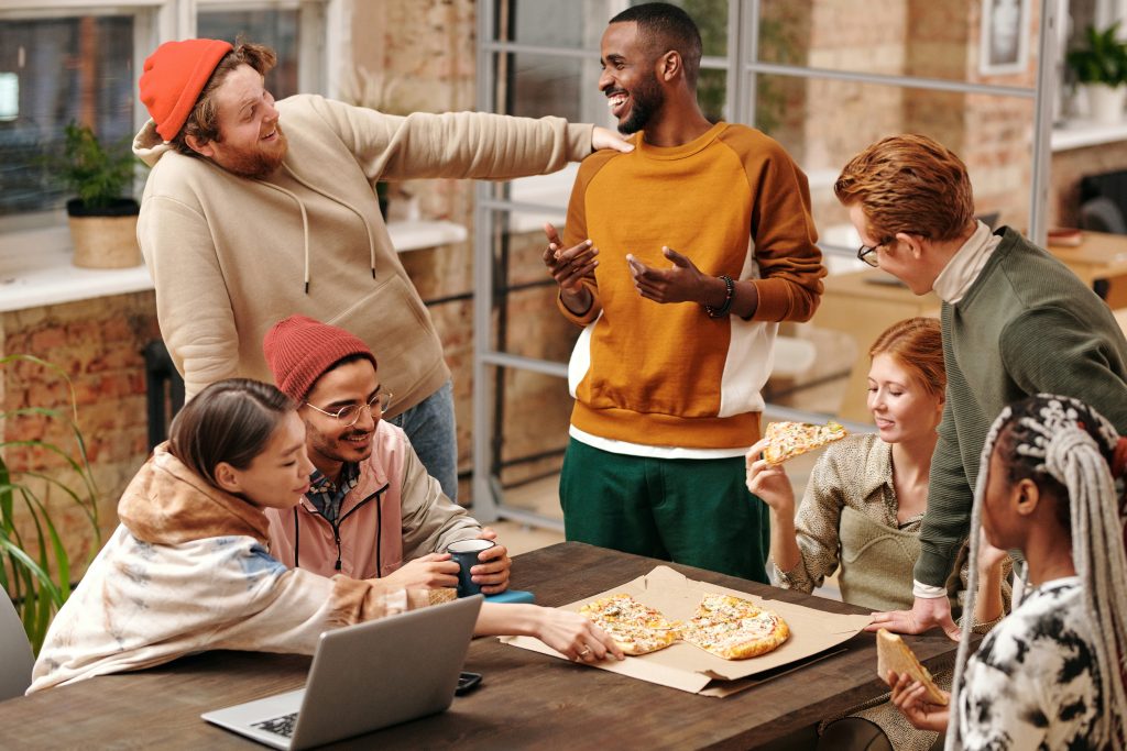 Group of friends sit at kitchen table laughing while eating pizza